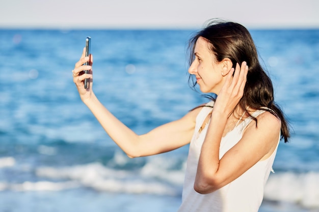 Junge kaukasische Frau im weißen Unterhemd macht Selfie am Strand nahe dem Meer in der Türkei auf ihrem Smartphone