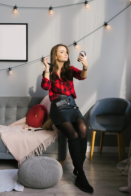 Junge kaukasische Frau in einem roten karierten Hemd und Rock lässt ihre Make-up-Farben mit einem Pinsel erröten, wenn sie zu Hause auf der Couch sitzt und in den Spiegel schaut