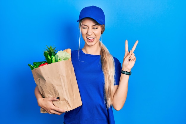 Junge kaukasische Frau in Kurieruniform mit Lebensmitteln aus dem Supermarkt, die mit glücklichem Gesicht lächelt und in die Kamera zwinkert und mit den Fingern Nummer zwei ein Siegeszeichen macht