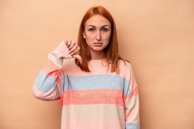 Junge kaukasische Frau isoliert auf beigem Hintergrund, die eine Abneigungsgeste mit dem Daumen nach unten zeigt Uneinigkeitskonzept