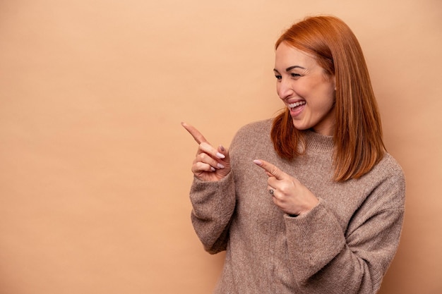 Junge kaukasische Frau isoliert auf beigem Hintergrund zeigt mit Daumenfinger weg lachend und sorglos