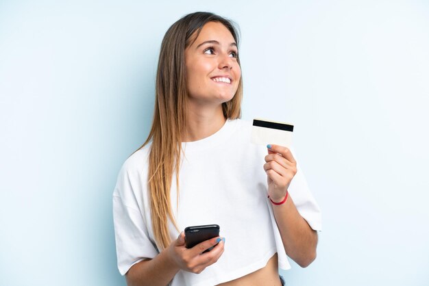 Junge kaukasische Frau isoliert auf blauem Hintergrund, die mit dem Handy mit einer Kreditkarte kauft, während sie nachdenkt
