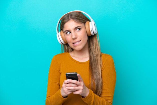 Junge kaukasische Frau isoliert auf blauem Hintergrund, die Musik mit einem Handy hört und denkt