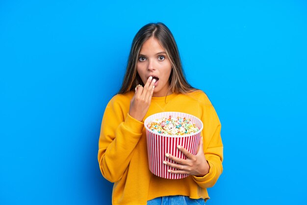 Junge kaukasische Frau isoliert auf blauem Hintergrund mit einem großen Eimer Popcorn