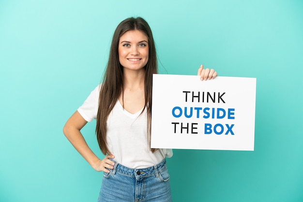 Junge kaukasische Frau isoliert auf blauem Hintergrund mit einem Plakat mit Text Think Outside The Box mit glücklichem Ausdruck