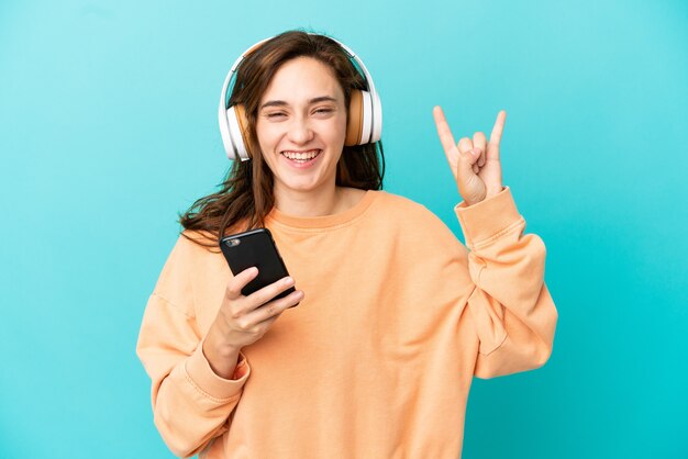 Junge kaukasische Frau isoliert auf blauem Hintergrund Musikhören mit einem Handy, das Rockgeste macht