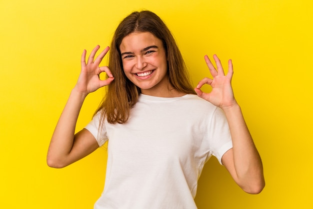 Junge kaukasische Frau isoliert auf gelbem Hintergrund fröhlich und selbstbewusst, die ok Geste zeigt