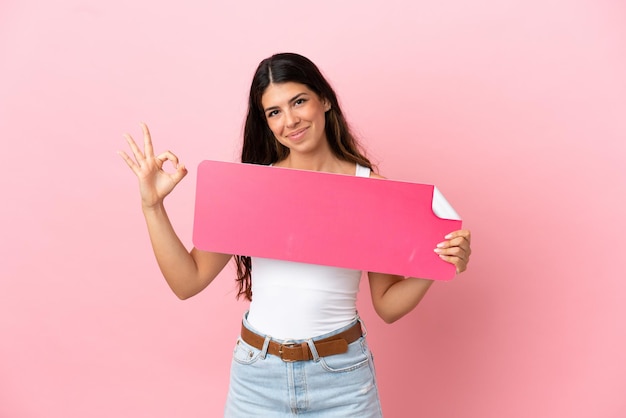 Junge kaukasische Frau isoliert auf rosa Hintergrund, die ein leeres Plakat hält und OK-Zeichen macht