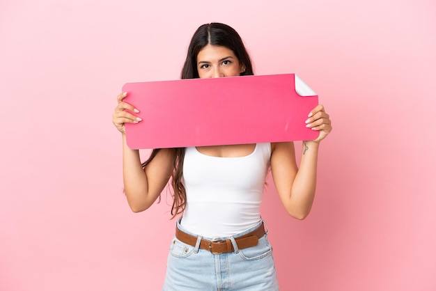 Junge kaukasische Frau isoliert auf rosa Hintergrund, die ein leeres Plakat hält und sich dahinter versteckt