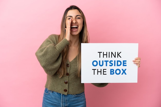 Junge kaukasische Frau isoliert auf rosa Hintergrund, die ein Plakat mit dem Text Think Outside The Box hält und schreit