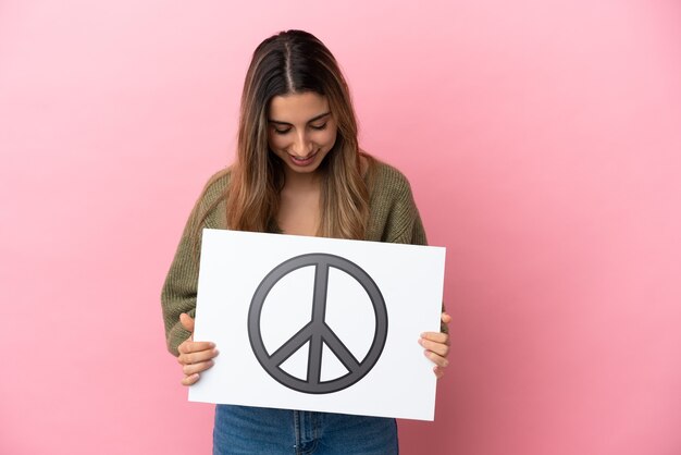 Junge kaukasische Frau isoliert auf rosa Hintergrund, die ein Plakat mit Friedenssymbol hält