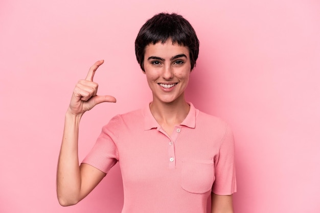 Junge kaukasische Frau isoliert auf rosa Hintergrund, die etwas Kleines mit Zeigefingern lächelnd und selbstbewusst hält