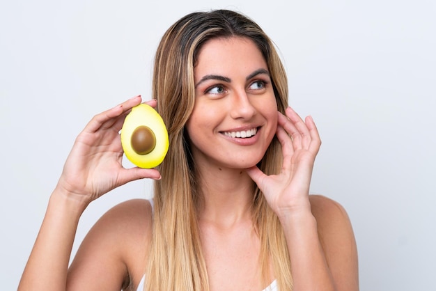 Junge kaukasische Frau isoliert auf weißem Hintergrund, die eine Avocado hält, während sie lächelt Close up Portrait