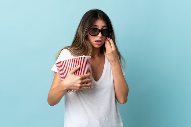 Junge kaukasische Frau lokalisiert auf Blau mit 3d Brille und hält einen großen Eimer Popcorn