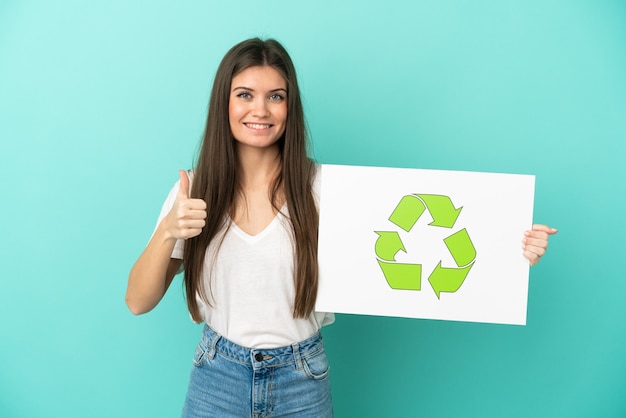 Junge kaukasische Frau lokalisiert auf blauem Hintergrund, der ein Plakat mit Recycling-Symbol mit Daumen nach oben hält