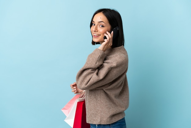 Junge kaukasische Frau lokalisiert auf blauem Hintergrund, der Einkaufstaschen hält und einen Freund mit ihrem Handy anruft