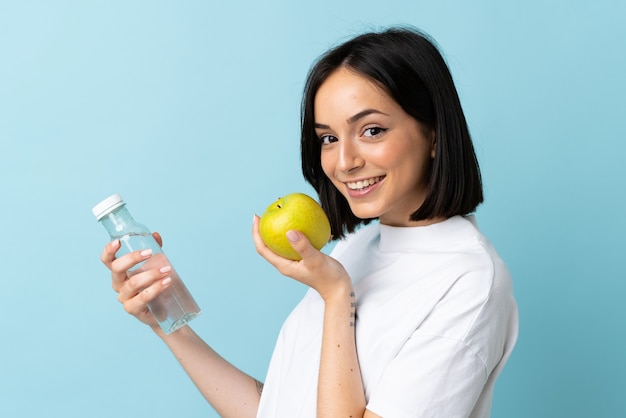 Junge kaukasische Frau lokalisiert auf blauem Hintergrund mit einem Apfel und mit einer Flasche Wasser