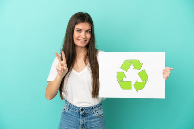 Junge kaukasische Frau lokalisiert auf blauer Wand, die ein Plakat mit Recycling-Ikone hält und einen Sieg feiert