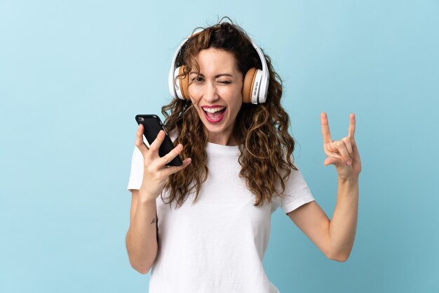 Junge kaukasische Frau lokalisiert auf blauer Wand, die Musik mit einem Handy hört, das Rockgeste macht