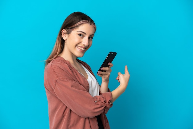 Junge kaukasische Frau lokalisiert auf blauer Wand unter Verwendung des Mobiltelefons und zurückweisend