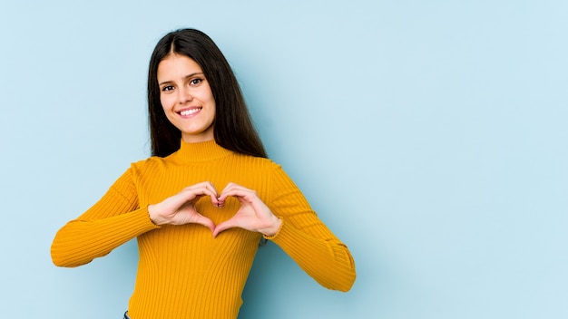 Junge kaukasische Frau lokalisiert auf der blauen Wand lächelnd und zeigt eine Herzform mit Händen.