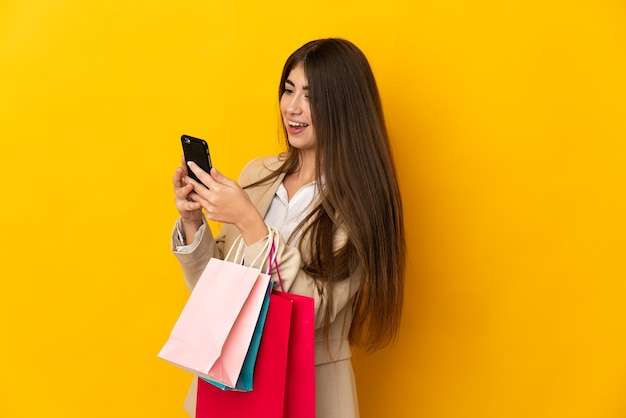Junge kaukasische Frau lokalisiert auf gelbem Hintergrund, der Einkaufstaschen hält und eine Nachricht mit ihrem Handy an einen Freund schreibt