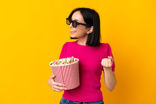 Junge kaukasische Frau lokalisiert auf gelbem Hintergrund mit 3D-Brille und hält einen großen Eimer Popcorn, während sie Seite schaut