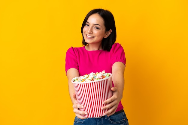 Junge kaukasische Frau lokalisiert auf gelber Wand, die einen großen Eimer Popcorn hält