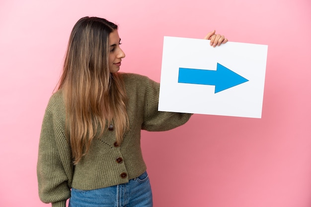 Junge kaukasische Frau lokalisiert auf rosa Hintergrund, der ein Plakat mit Pfeilsymbol hält