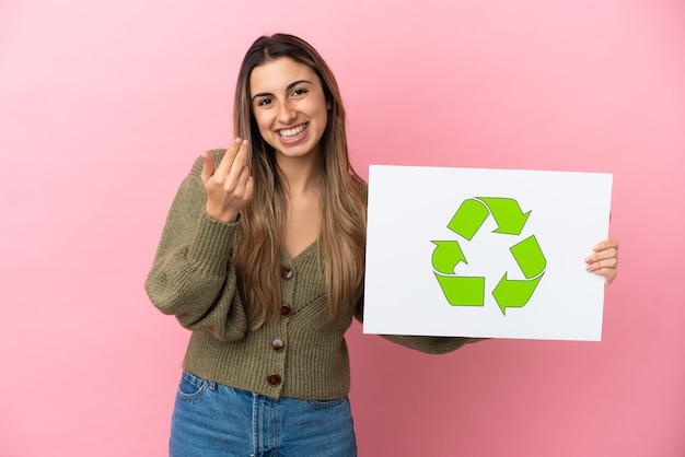 Junge kaukasische Frau lokalisiert auf rosa Hintergrund, der ein Plakat mit Recycling-Symbol hält und kommende Geste tut