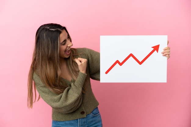 Junge kaukasische Frau lokalisiert auf rosa Hintergrund, der ein Zeichen mit einem wachsenden Statistikpfeilsymbol hält und einen Sieg feiert