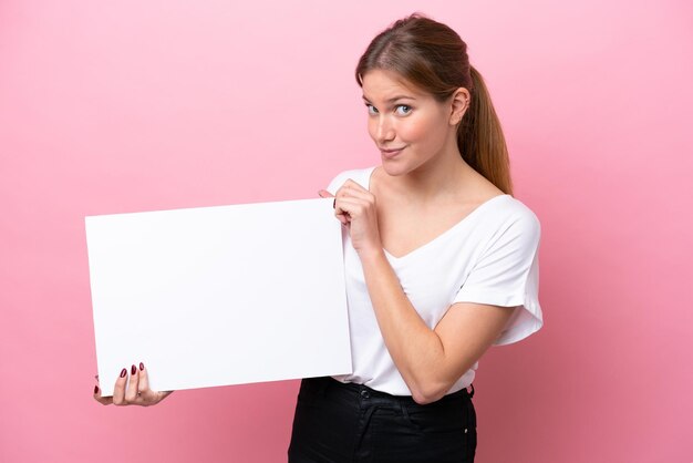 Junge kaukasische Frau lokalisiert auf rosa Hintergrund, die ein leeres Plakat mit glücklichem Ausdruck hält