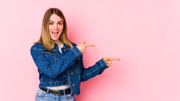 Junge kaukasische Frau lokalisiert auf rosa Wand aufgeregt, die mit Zeigefingern weg zeigt.