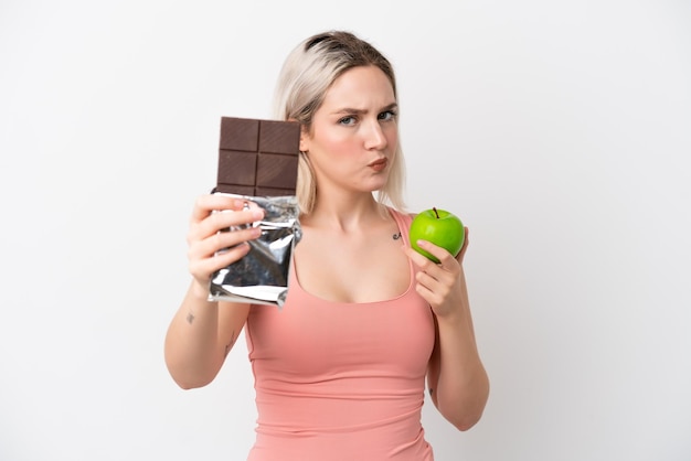 Junge kaukasische Frau lokalisiert auf weißem Hintergrund, der eine Schokoladentafel in einer Hand und einen Apfel in der anderen nimmt