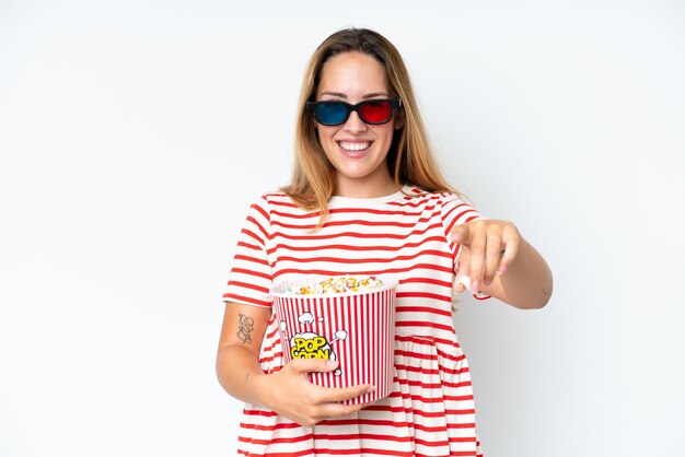 Junge kaukasische Frau lokalisiert auf weißem Hintergrund mit 3D-Brille und hält einen großen Eimer Popcorn, während sie nach vorne zeigt