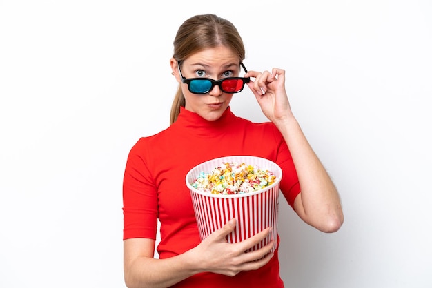 Junge kaukasische Frau lokalisiert auf weißem Hintergrund überrascht mit 3D-Brille und hält einen großen Eimer Popcorn