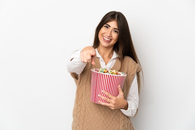 Junge kaukasische Frau lokalisiert auf weißer Wand, die einen großen Eimer Popcorn hält, während sie nach vorne zeigt