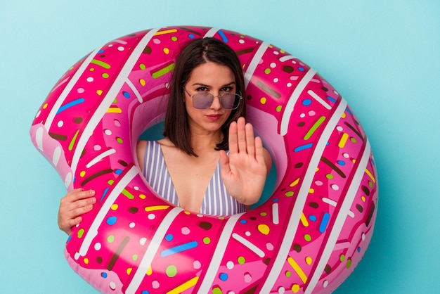 Junge kaukasische Frau mit aufblasbarem Donut isoliert auf blauem Hintergrund, die mit ausgestreckter Hand steht und ein Stoppschild zeigt, das Sie daran hindert