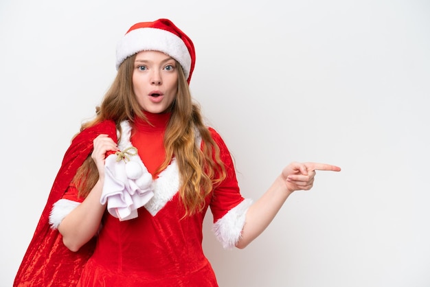 Junge kaukasische Frau mit dem Weihnachtskleid, das den Weihnachtssack lokalisiert auf weißem Hintergrund hält, überrascht und zeigt Seite