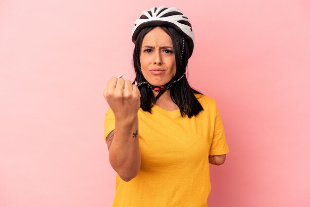 Junge kaukasische Frau mit einem Arm, der einen Fahrradhelm trägt, der auf rosafarbenem Hintergrund isoliert ist und die Faust zur Kamera zeigt, aggressiver Gesichtsausdruck.