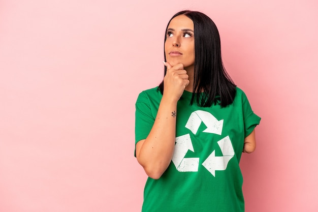 Junge kaukasische Frau mit einem Arm recycelten Abfall einzeln auf rosafarbenem Hintergrund, der seitlich mit zweifelhaftem und skeptischem Ausdruck schaut.