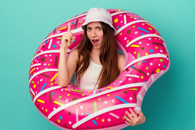 Junge kaukasische Frau mit einem aufblasbaren Donut isoliert auf blauer Wand mit einer Idee, Inspirationskonzept.