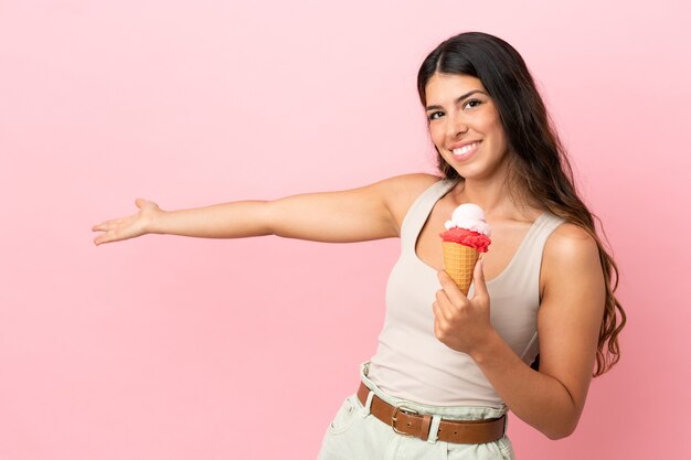 Junge kaukasische Frau mit einem Kornett-Eis isoliert auf rosa Hintergrund, das die Hände zur Seite ausstreckt, um zum Kommen einzuladen?