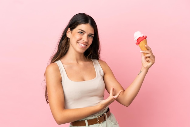 Junge kaukasische Frau mit einem Kornett-Eis isoliert auf rosa Hintergrund, das die Hände zur Seite ausstreckt, um zum Kommen einzuladen?