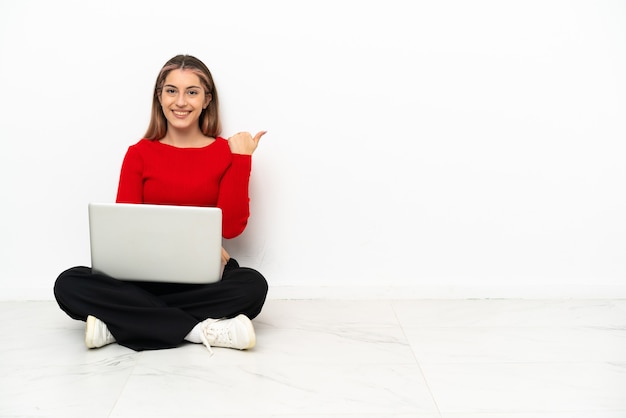 Junge kaukasische Frau mit einem Laptop, der auf dem Boden sitzt, der zur Seite zeigt, um ein Produkt zu präsentieren