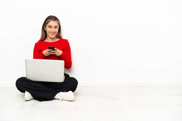 Junge kaukasische Frau mit einem Laptop, der auf dem Boden sitzt und eine Nachricht mit dem Handy sendet