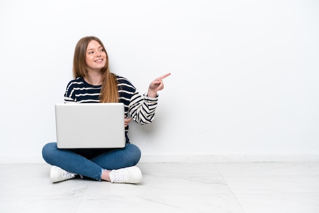 Junge kaukasische Frau mit einem Laptop, der isoliert auf weißem Hintergrund auf dem Boden sitzt, mit dem Finger zur Seite zeigt und ein Produkt präsentiert