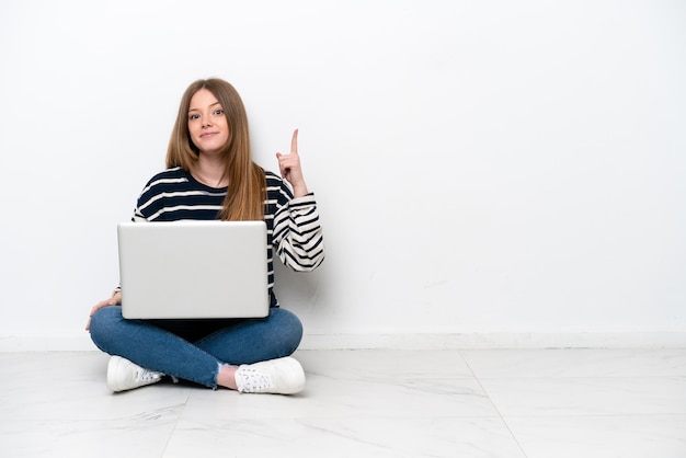 Junge kaukasische Frau mit einem Laptop, der isoliert auf weißem Hintergrund auf dem Boden sitzt und mit dem Zeigefinger auf eine großartige Idee zeigt