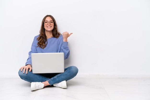 Junge kaukasische Frau mit einem Laptop, der isoliert auf weißem Hintergrund auf dem Boden sitzt und zur Seite zeigt, um ein Produkt zu präsentieren