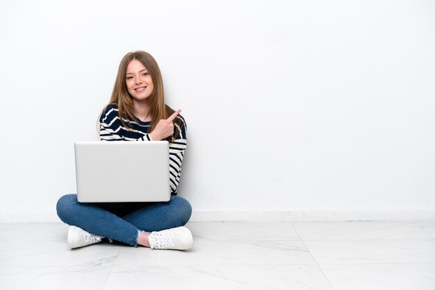 Junge kaukasische Frau mit einem Laptop, der isoliert auf weißem Hintergrund auf dem Boden sitzt und zur Seite zeigt, um ein Produkt zu präsentieren